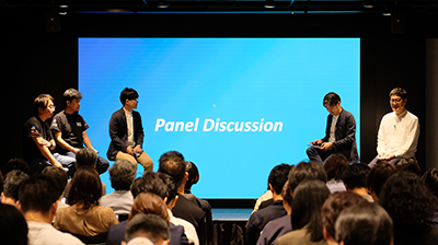 セミナー当日の写真。「Panel Discussion」と書かれたプロジェクタースクリーンを背景に、5名のパネリストが左右二手に分かれて座っている。