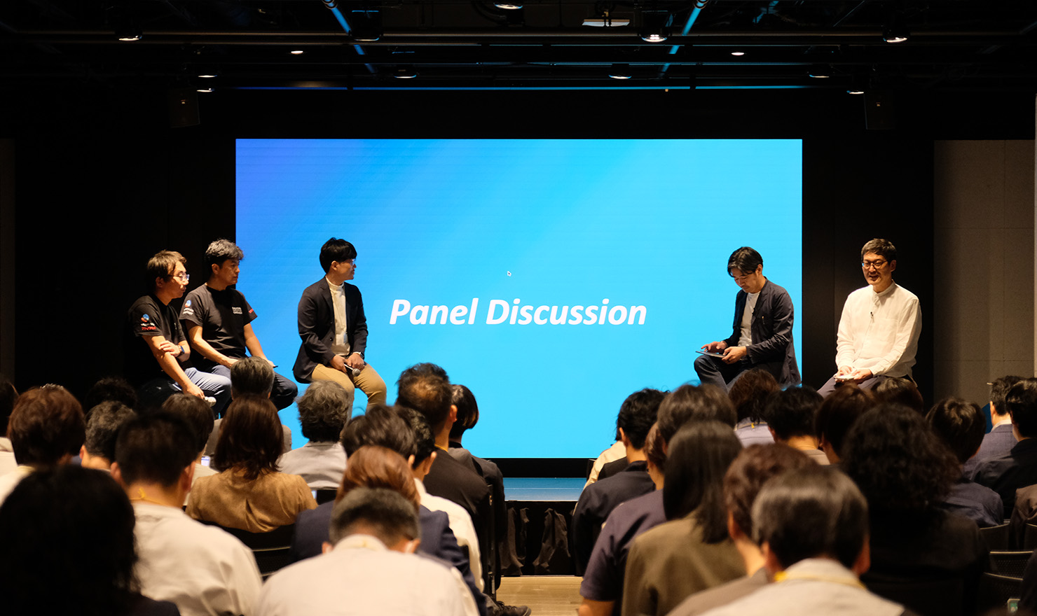 セミナー当日の写真。「Panel Discussion」と書かれたプロジェクタースクリーンを背景に、5名のパネリストが左右二手に分かれて座っている。